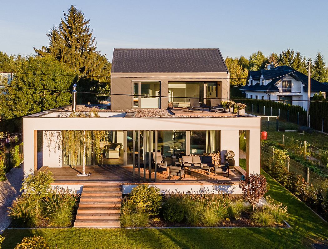 A modern, glass-walled house nestled in a lush, forested landscape, with a large deck and wooden stairs leading to the ground.