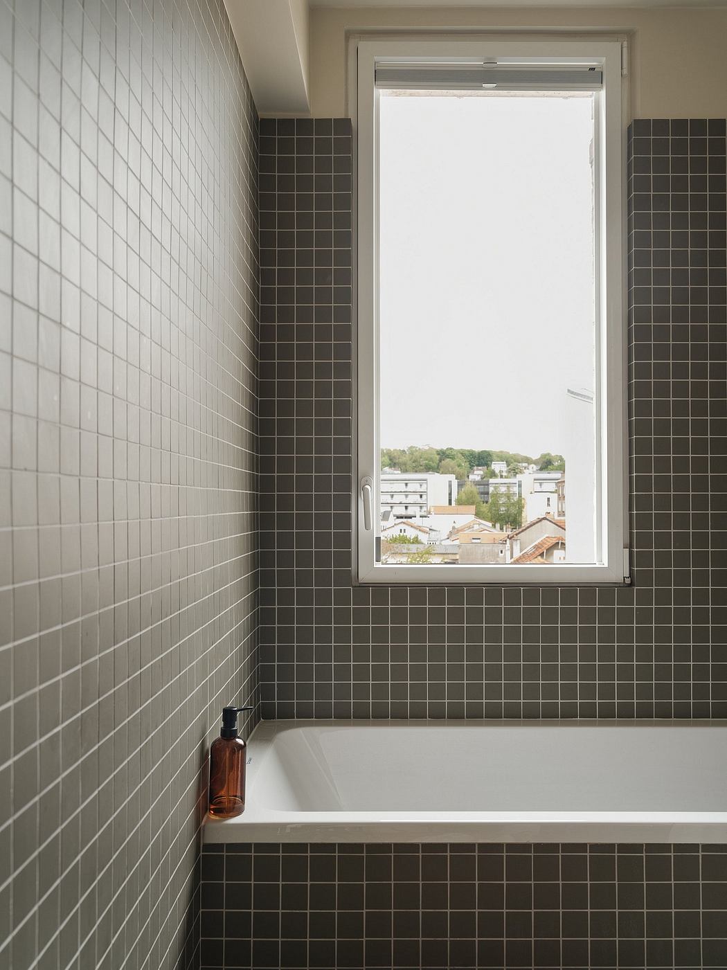 A modern bathroom with dark tile walls, a window overlooking a cityscape, and a minimalist bathtub.