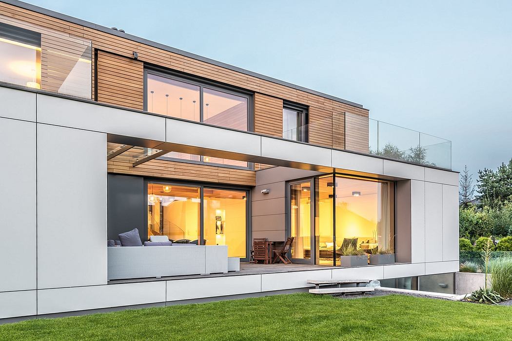 A modern, glass-enclosed house with wooden accents, surrounded by a grassy lawn and vegetation.