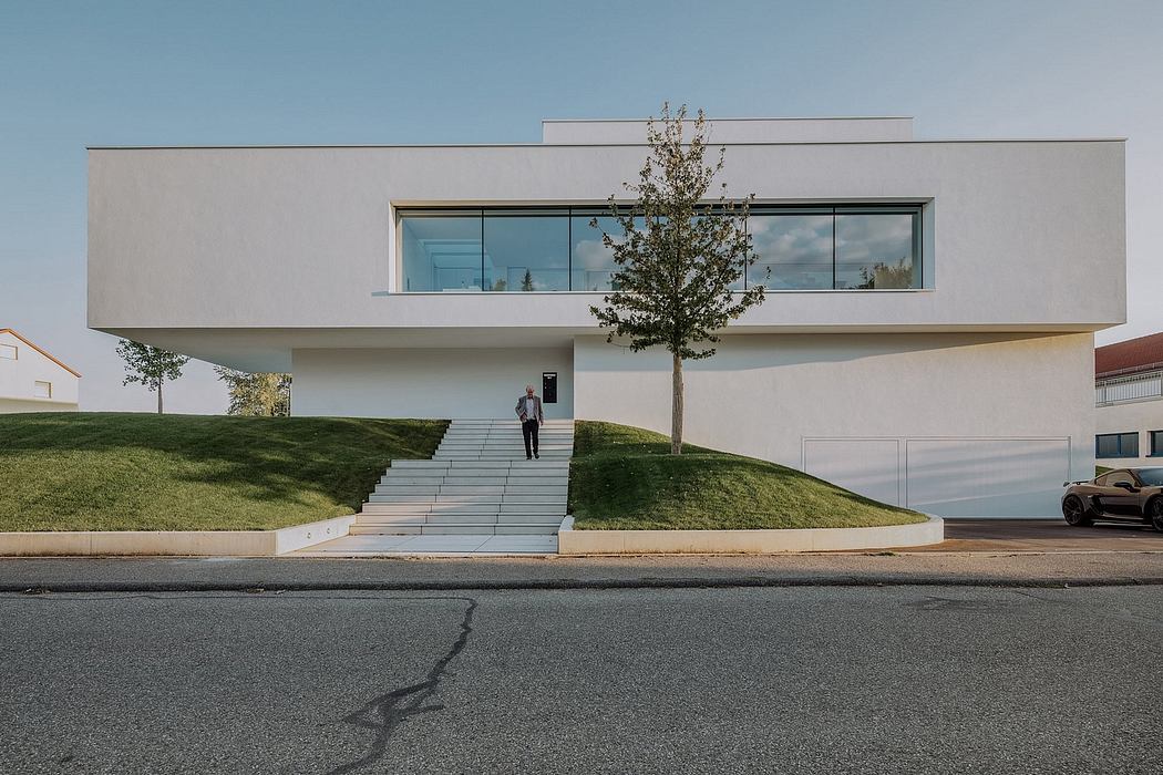 A modern, minimalist building with large glass windows, a tree in the foreground, and stairs leading up to the entrance.