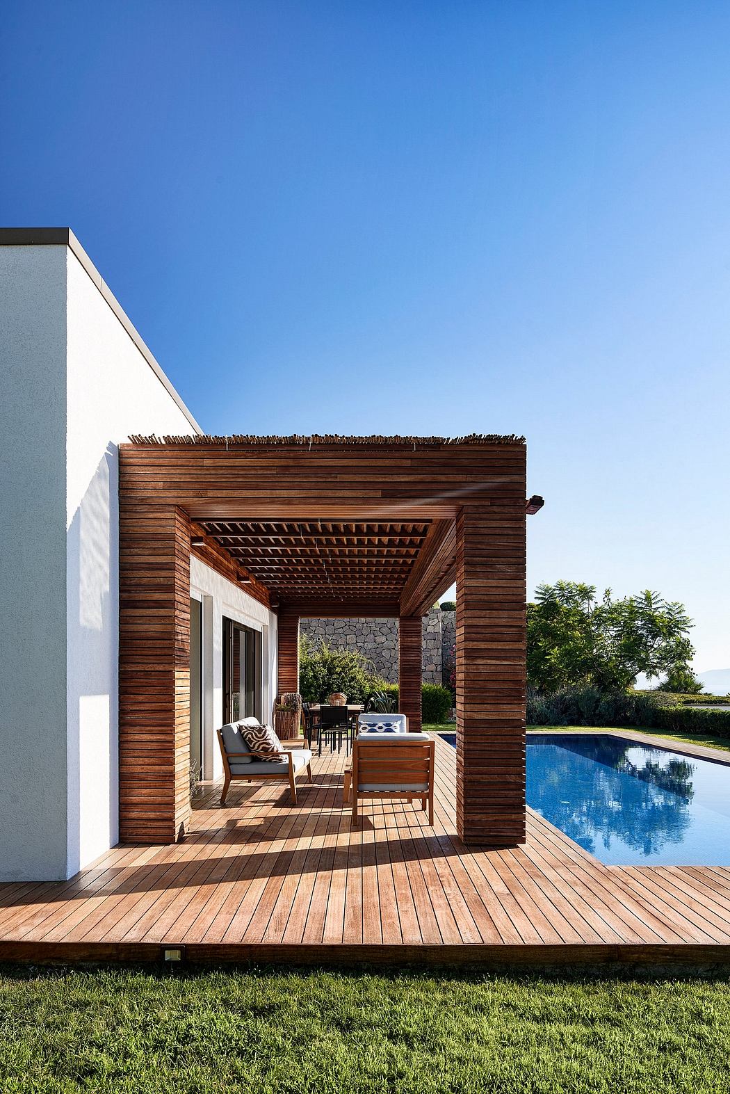 A modern wooden pergola with a stone wall and a rectangular pool, surrounded by lush greenery.