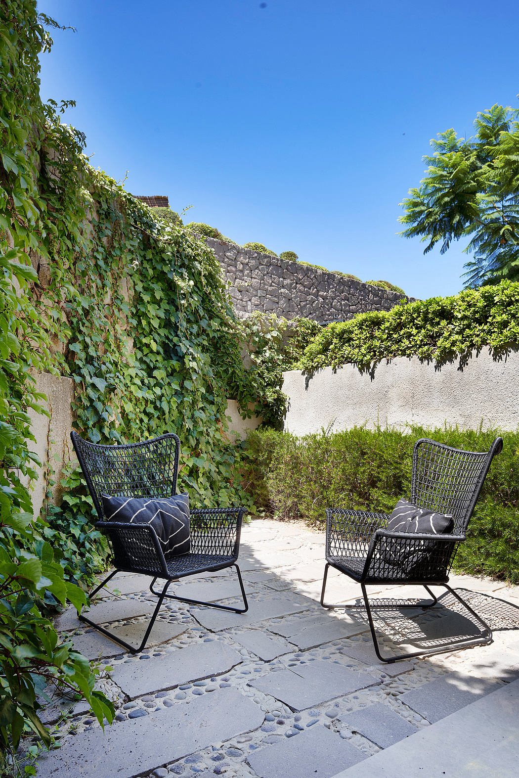 Secluded garden patio with stone walls, lush greenery, and modern wicker furniture.