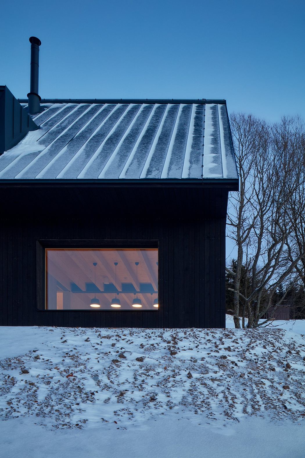A dark wooden cabin with a snow-covered roof and a large window illuminating the interior.