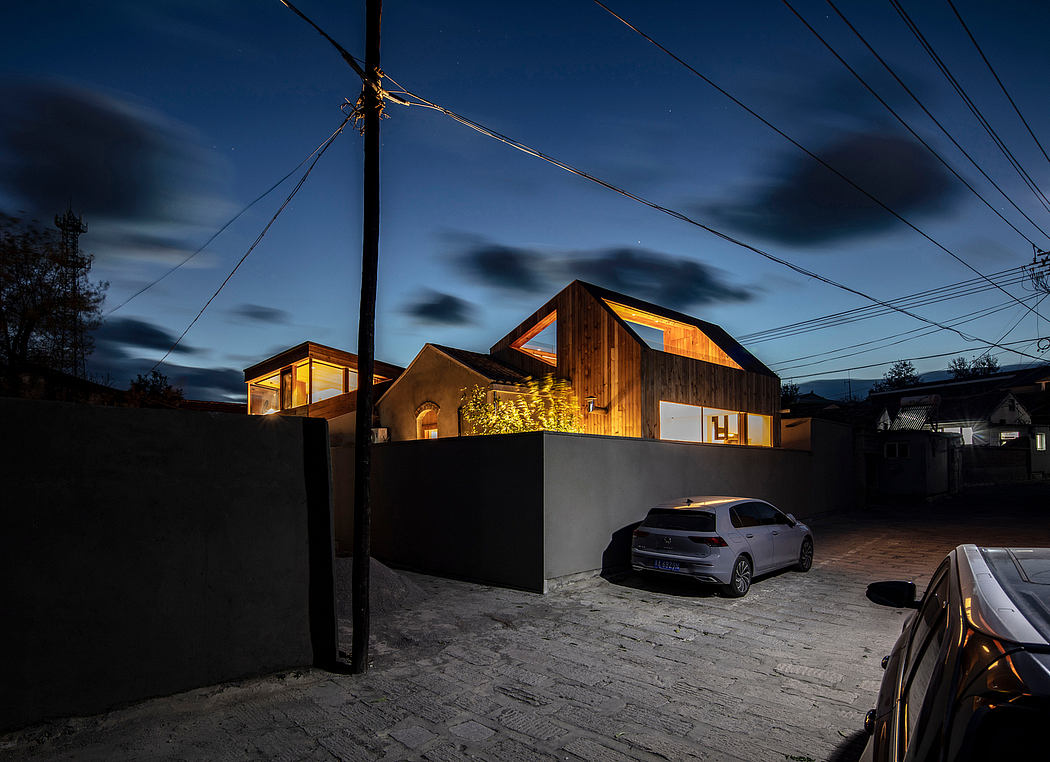 Modern wooden house with illuminated windows and parked cars on a cobblestone driveway at night.
