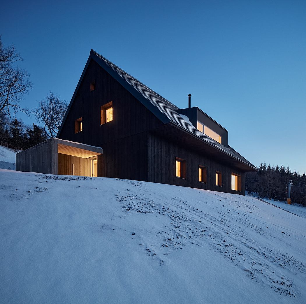 A modern, dark-colored wooden cabin nestled in a snowy landscape, with glowing windows.