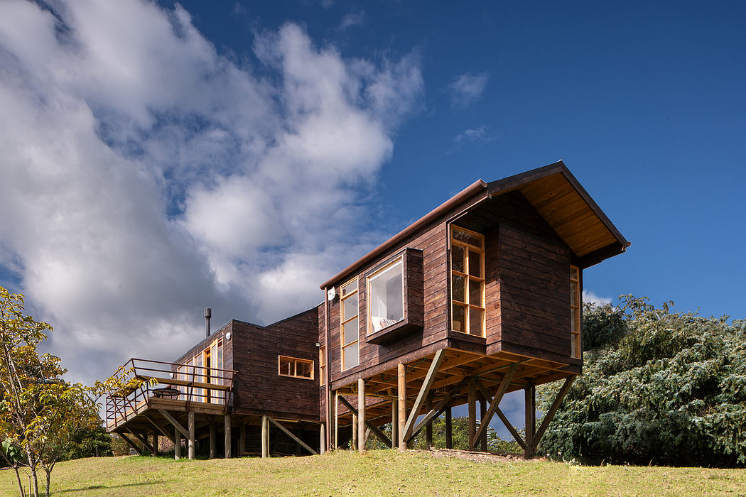 A distinctive wooden cabin elevated on stilts, with a covered porch and large windows against a cloudy sky.