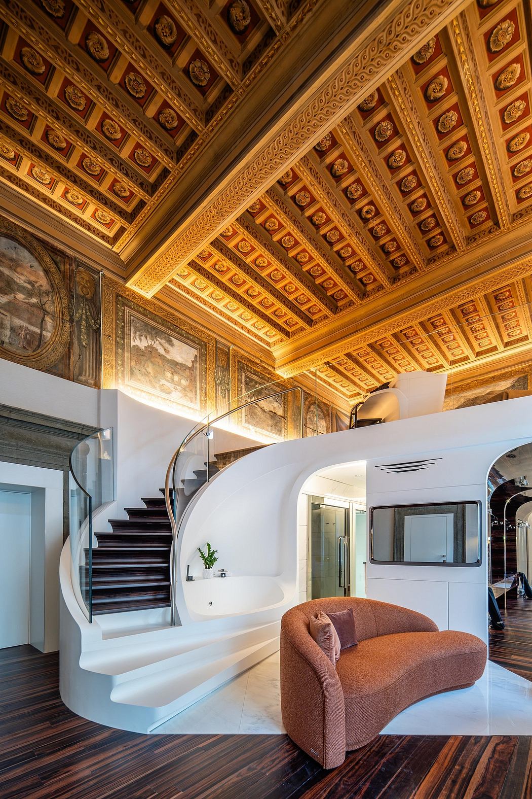 Ornate, gilded ceiling contrasts with modern, curving staircase and plush seating.