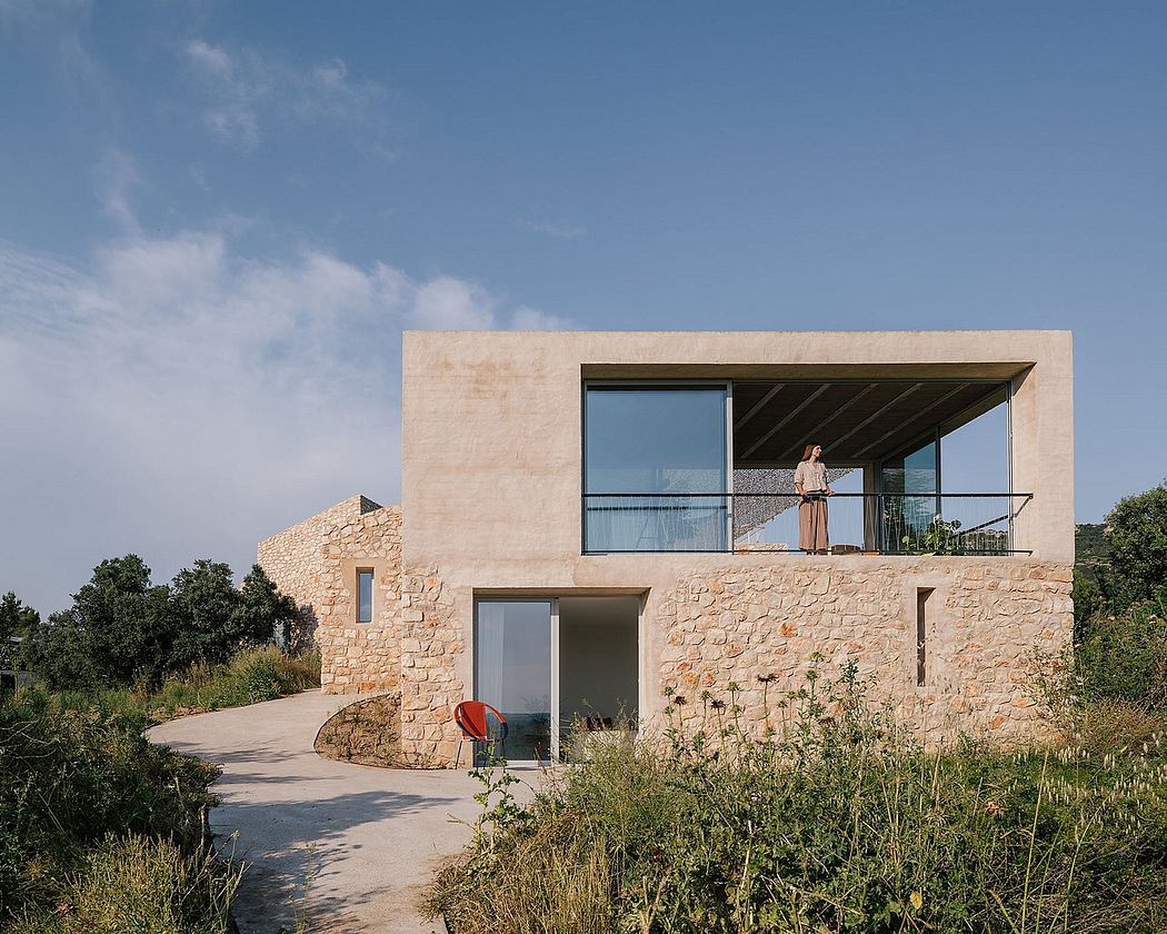 A modern stone house with large windows, a balcony, and a lush natural landscape.