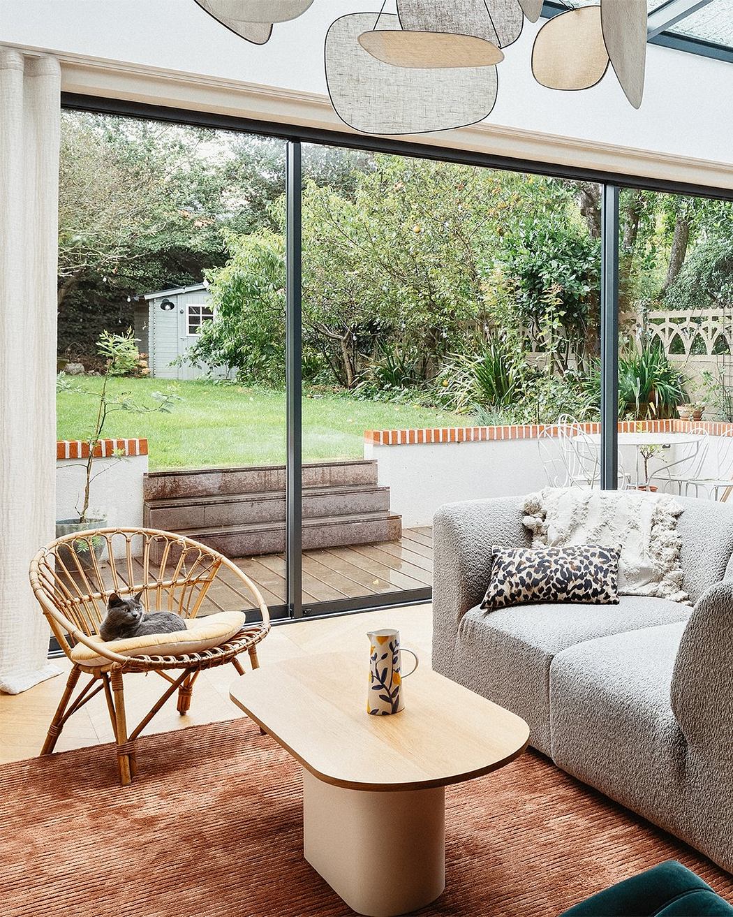 Modern living room with sleek furniture, wide windows overlooking lush greenery, and a cozy cat perched in the rattan chair.