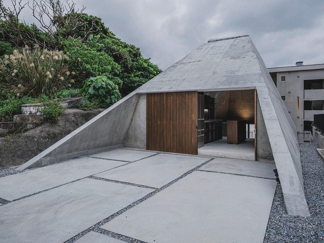 A modern, triangular concrete structure with a wooden entrance and surrounding greenery.