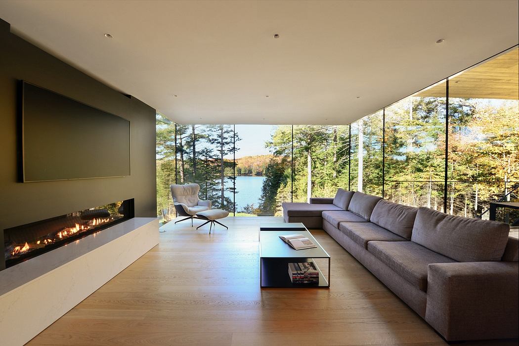 A modern, open-concept living room with a floor-to-ceiling glass wall overlooking a scenic lake and forest.