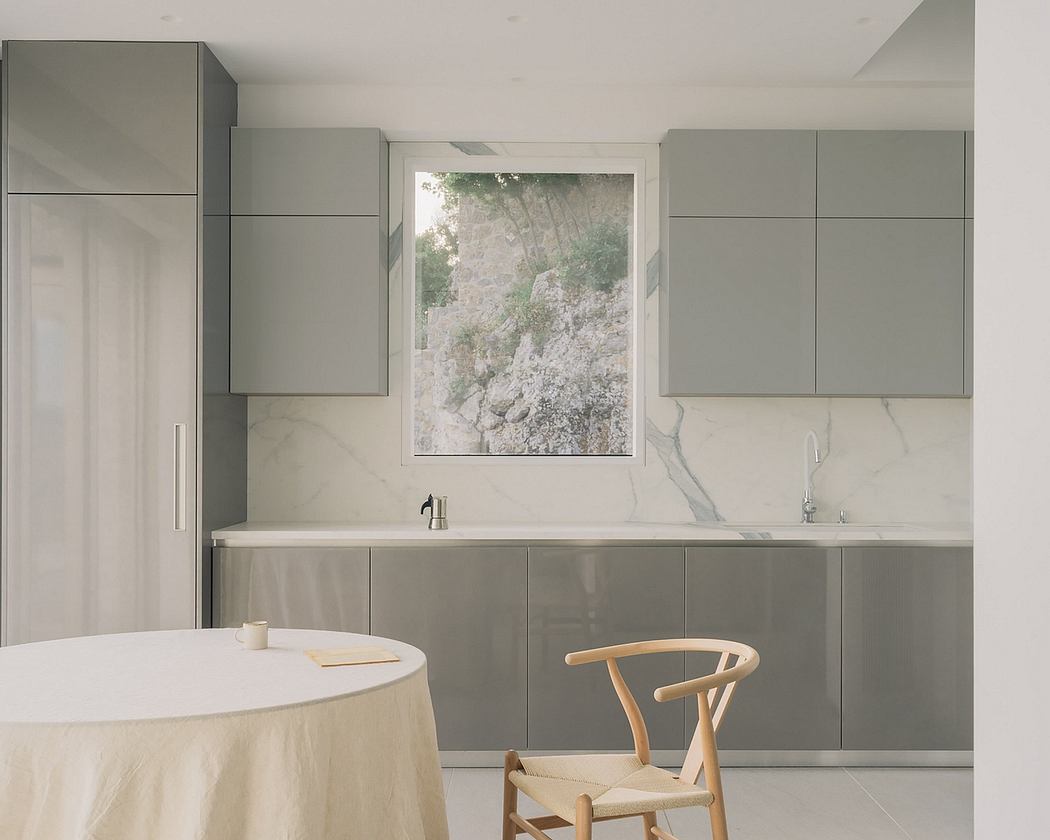 Modern kitchen with sleek gray cabinetry, marble countertops, and a large window overlooking nature.