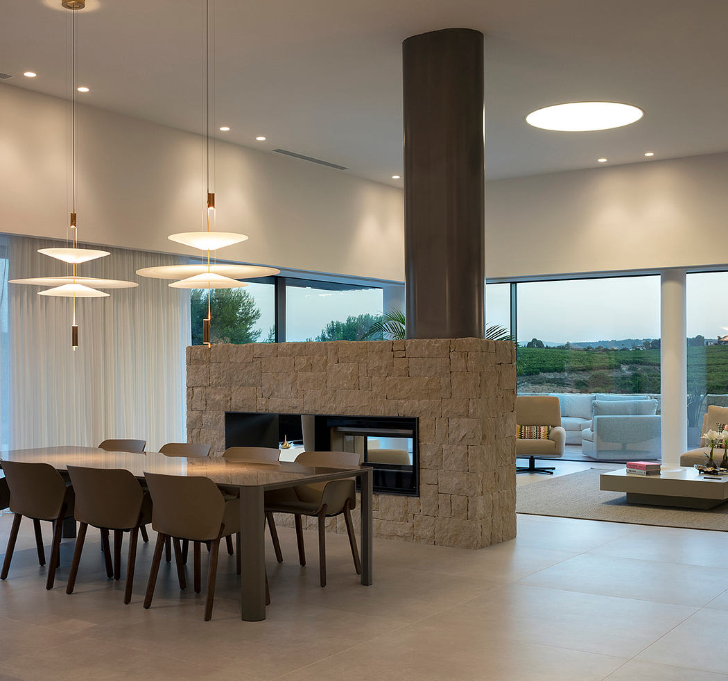 A modern dining area with a large stone fireplace, pendant lights, and floor-to-ceiling windows.