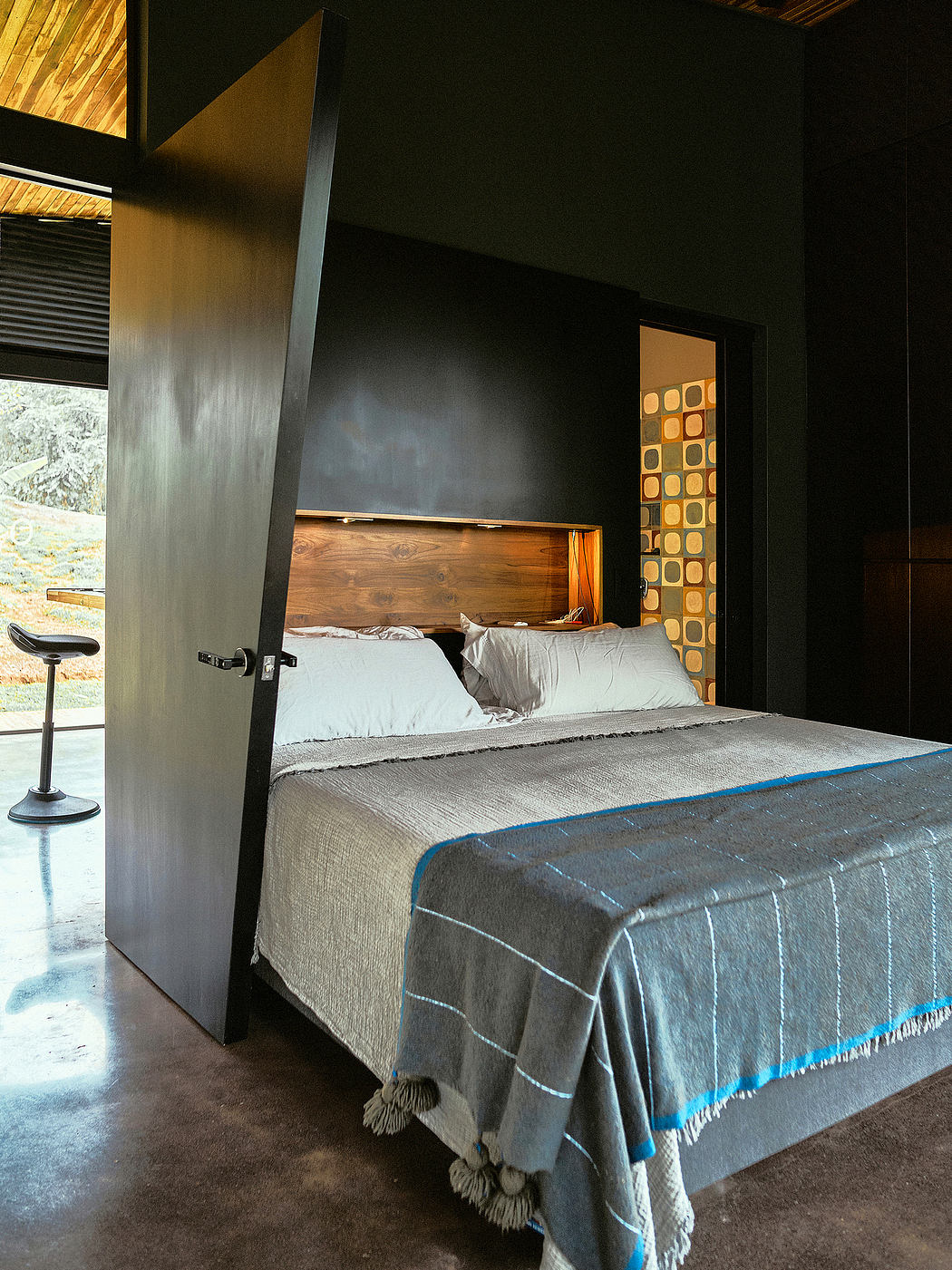 A modern, minimalist bedroom with a wooden headboard, patterned accent wall, and tasseled bedding.