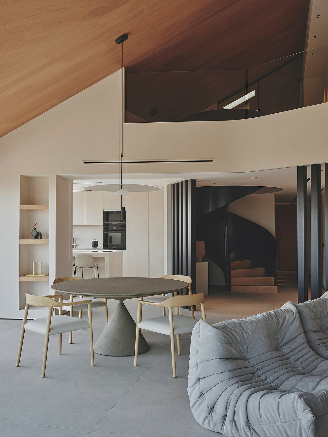 A modern open-plan living space with a round dining table, cozy sofa, and spiral staircase.