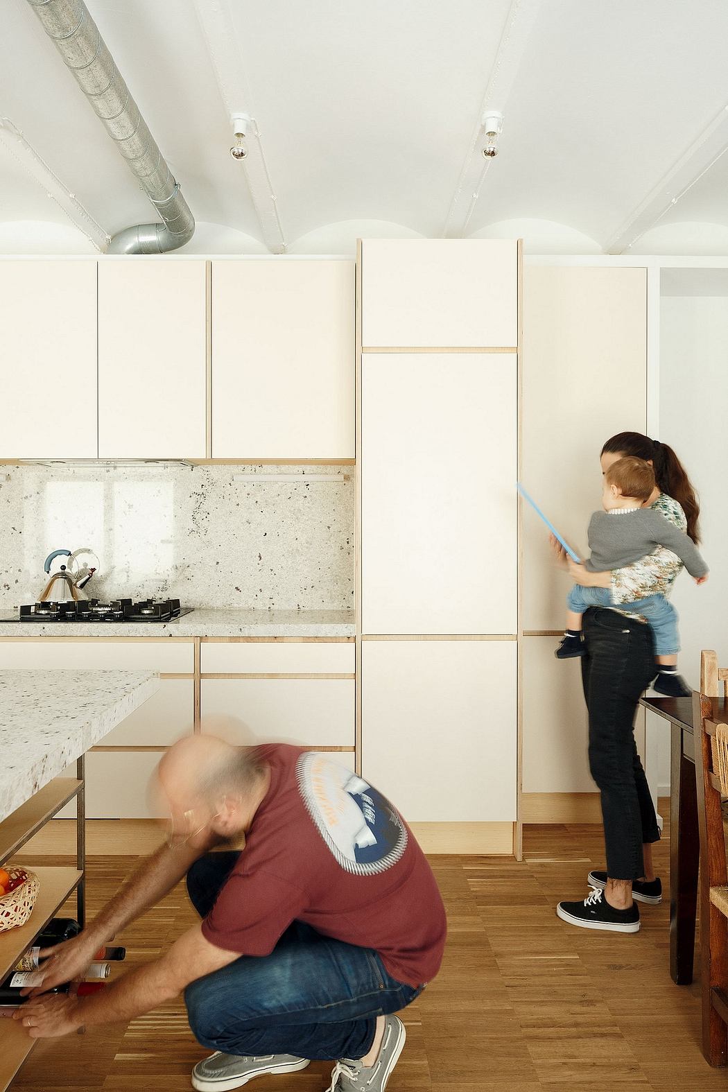 Modern kitchen with minimalist cabinetry, concrete backsplash, and people working on various tasks.