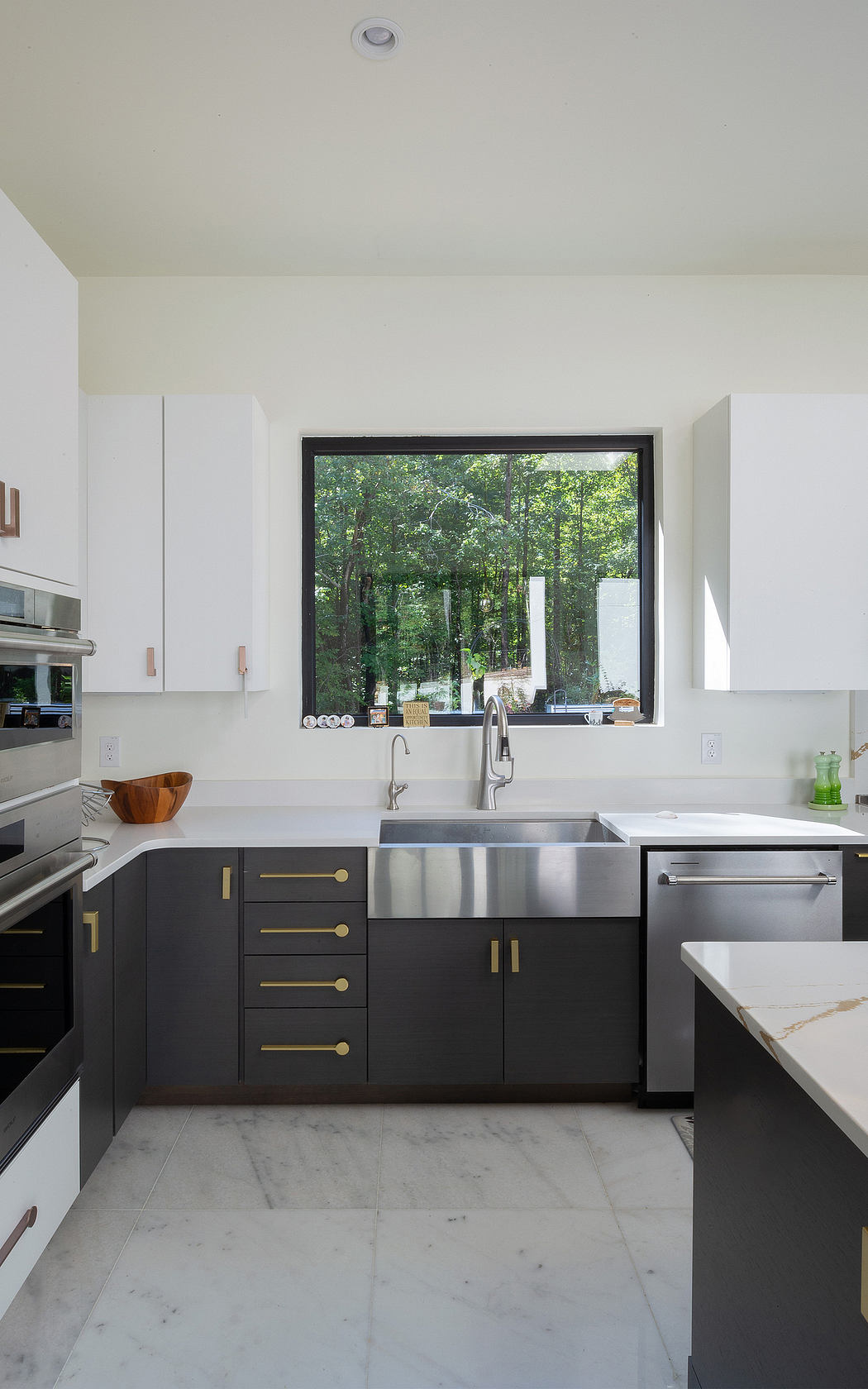 A modern kitchen with dark cabinetry, stainless steel appliances, and a large window overlooking a wooded area.