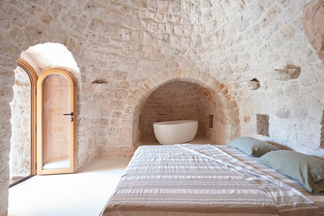 Rustic stone arched alcove with a modern freestanding bathtub, hardwood floors, and soft bedding.
