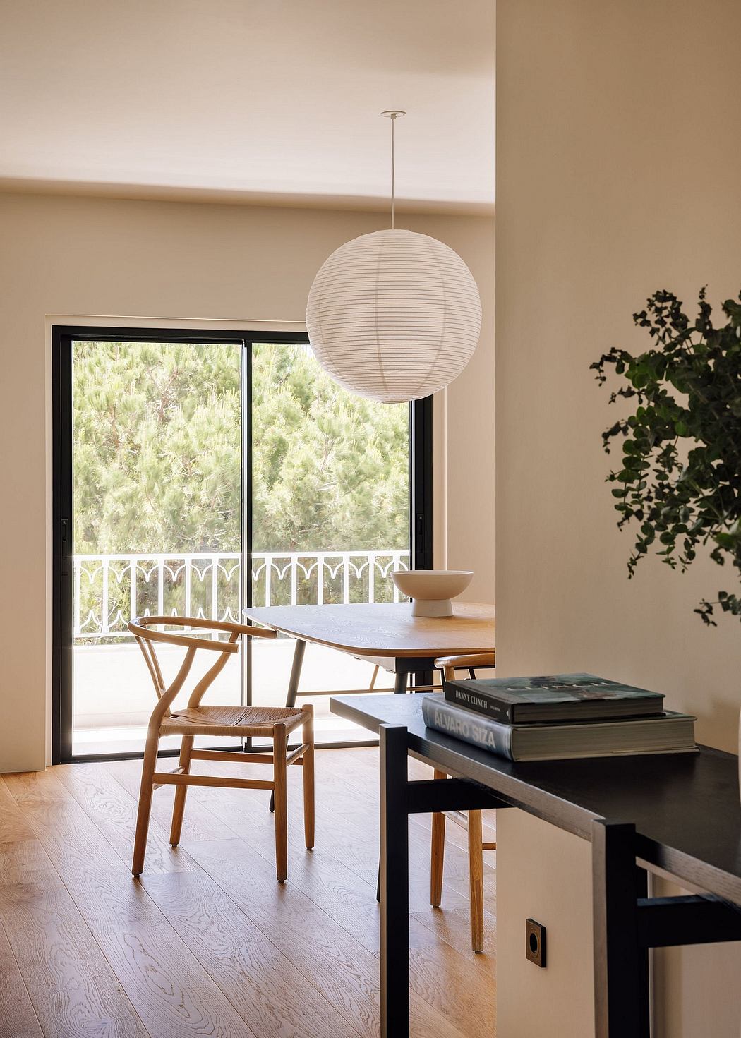 Minimalist dining area with modern lighting, wooden chairs, and a plant against scenic window.