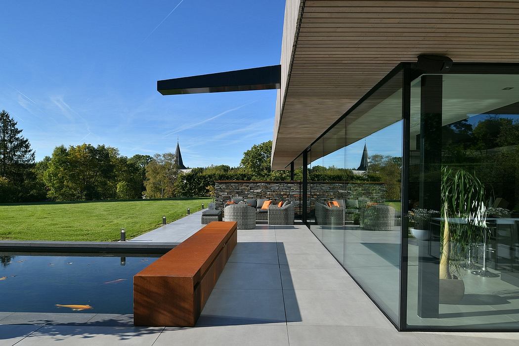 Modern glass-walled pavilion with an outdoor living area, reflecting pool, and surrounding lush greenery.