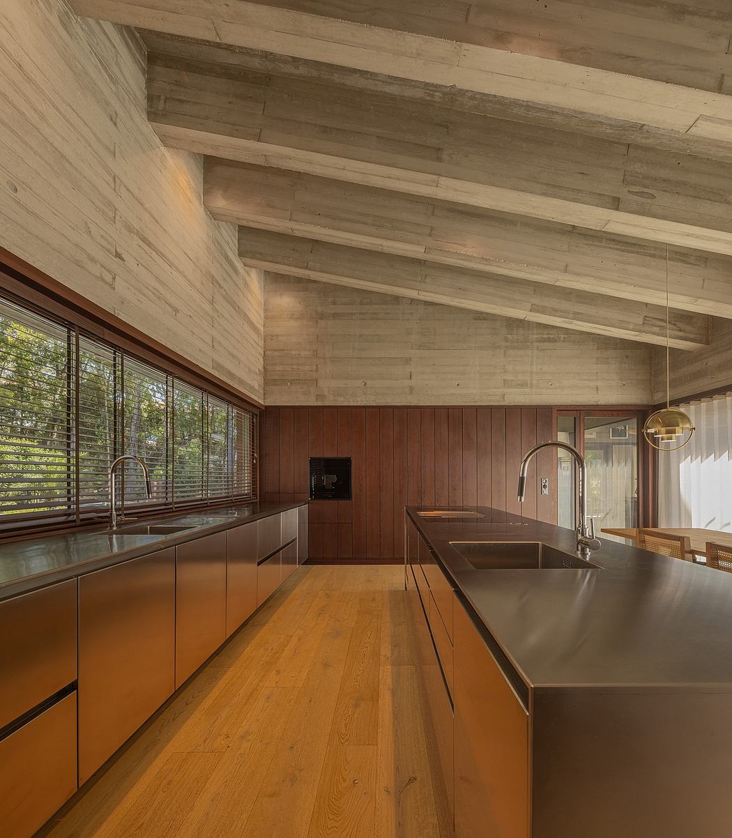 A modern open-plan kitchen with warm wood paneled walls and ceilings, stainless steel countertops, and large windows.
