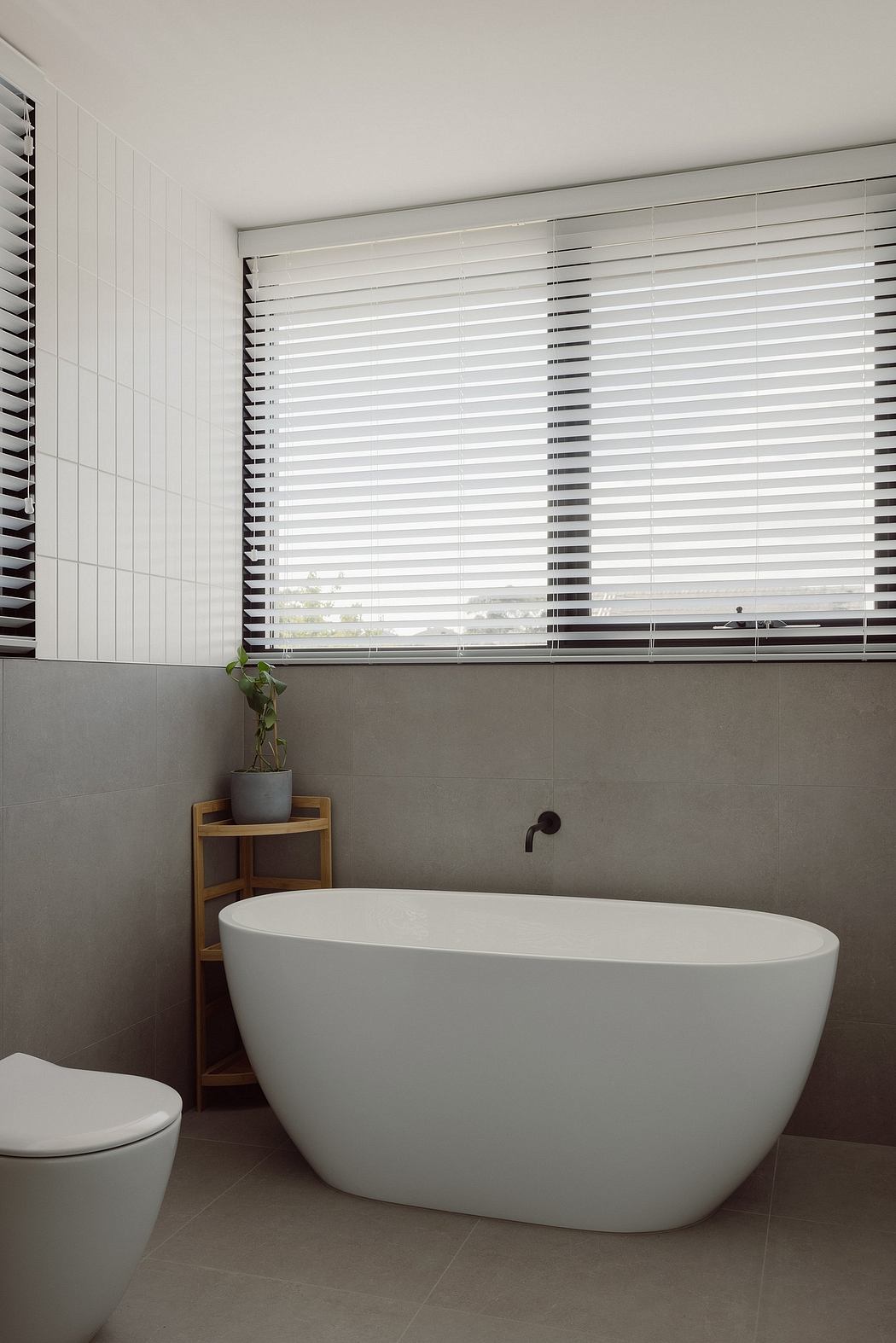 Sleek white freestanding tub, minimalist blinds, and a potted plant accent the modern bathroom.