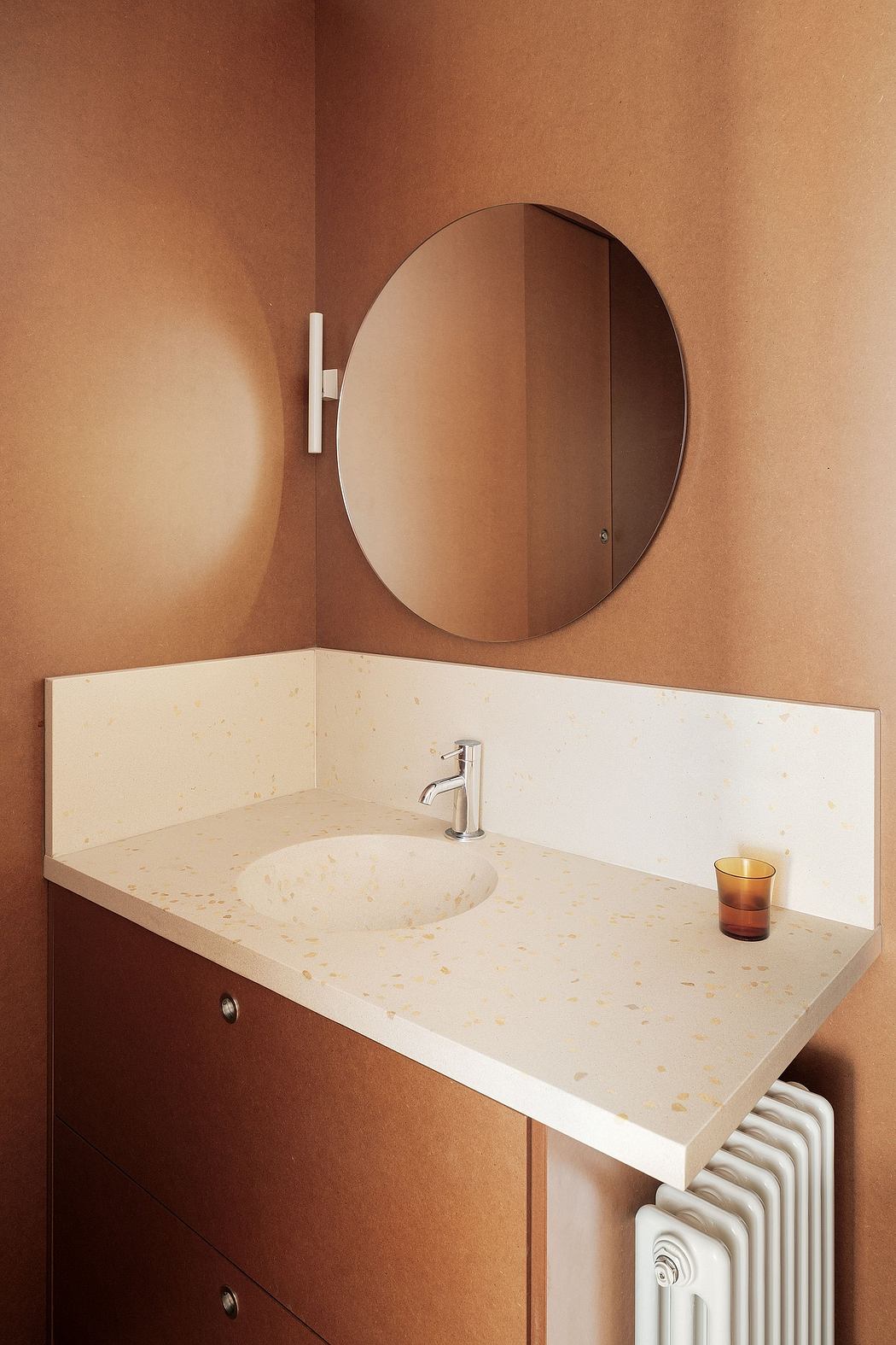 A circular mirror above a white countertop with a faucet and amber glass vase.