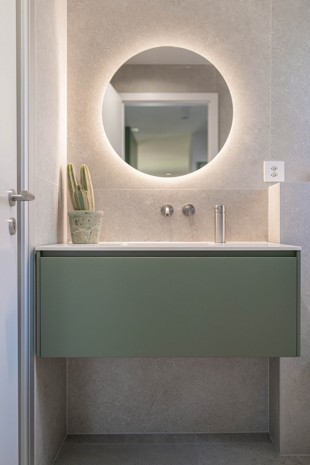 Minimalist bathroom with a circular backlit mirror, green vanity, and a potted cactus.