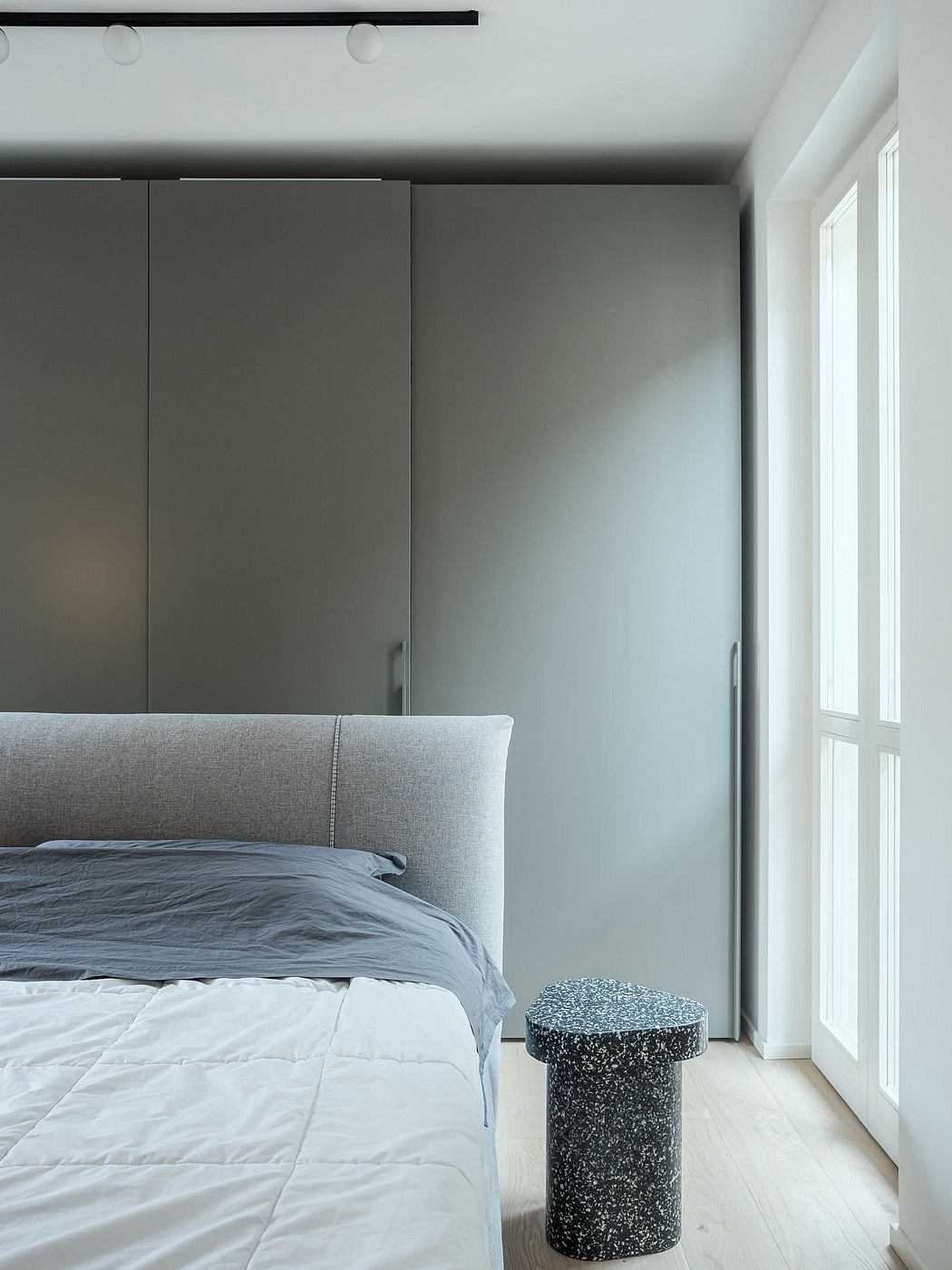 A minimalist bedroom with gray walls, a large window, and a sleek, gray upholstered bed.