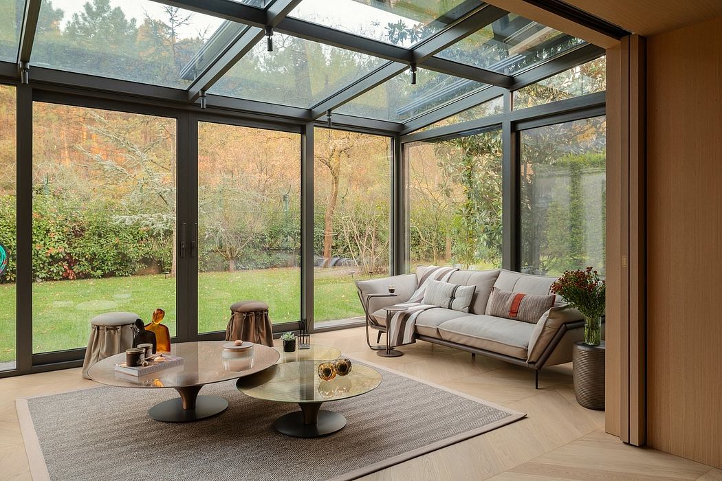 A cozy sunroom with sleek glass walls, a plush sofa, and modern glass coffee tables overlooking a lush garden.