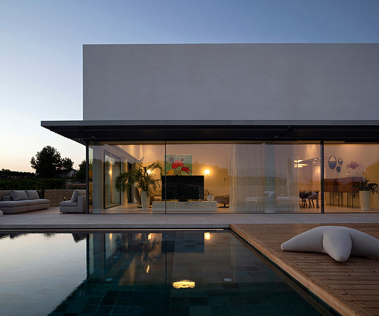 Modern minimalist house with sleek glass walls, pool, and deck at sunset.