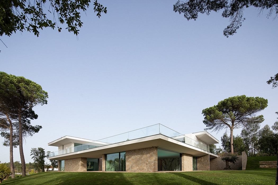Modern, glass-walled house nestled in a lush, wooded landscape with mature pine trees.