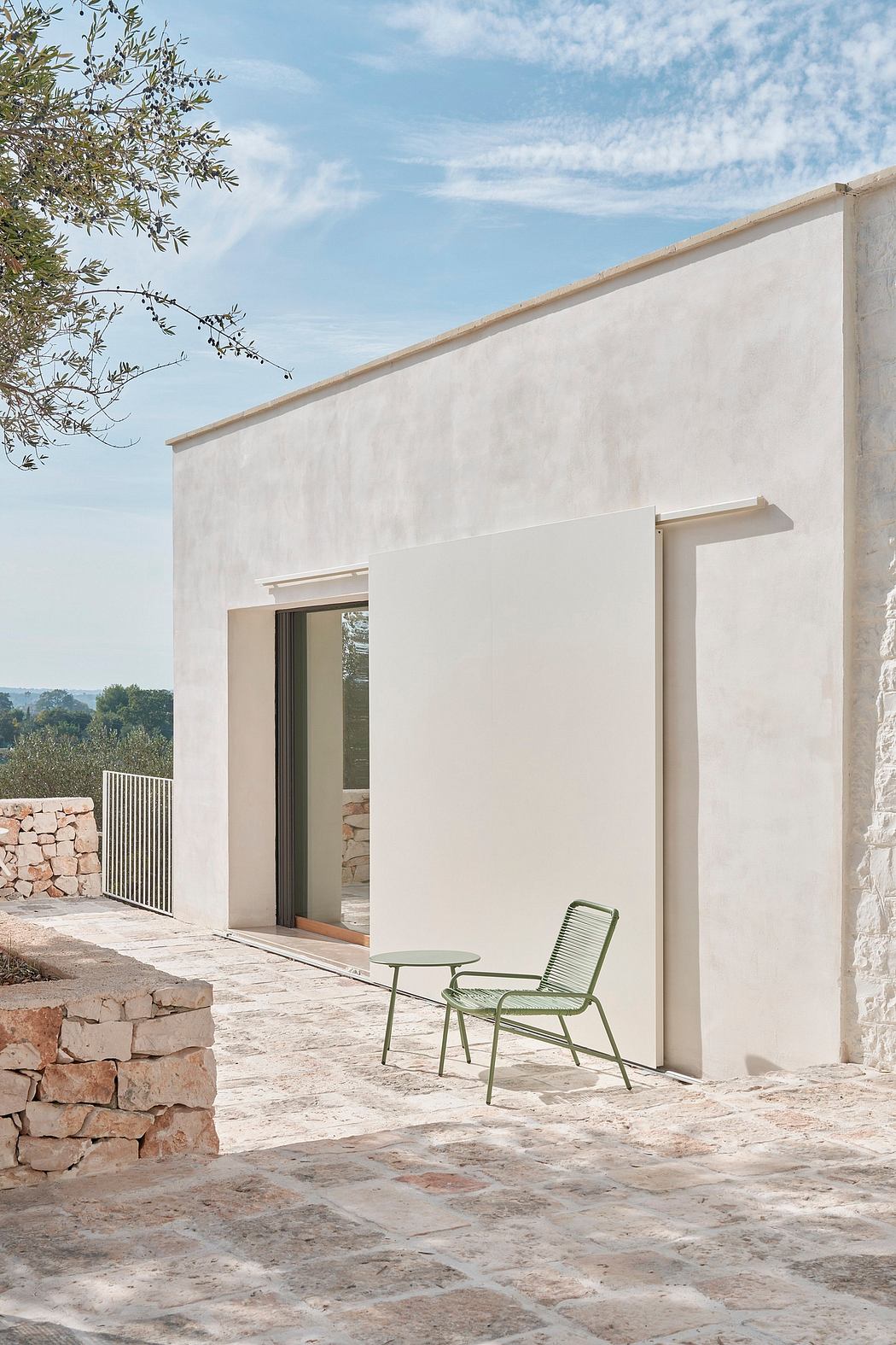 A minimalist exterior with clean white walls, stone paving, and a green metal chair inviting relaxation.