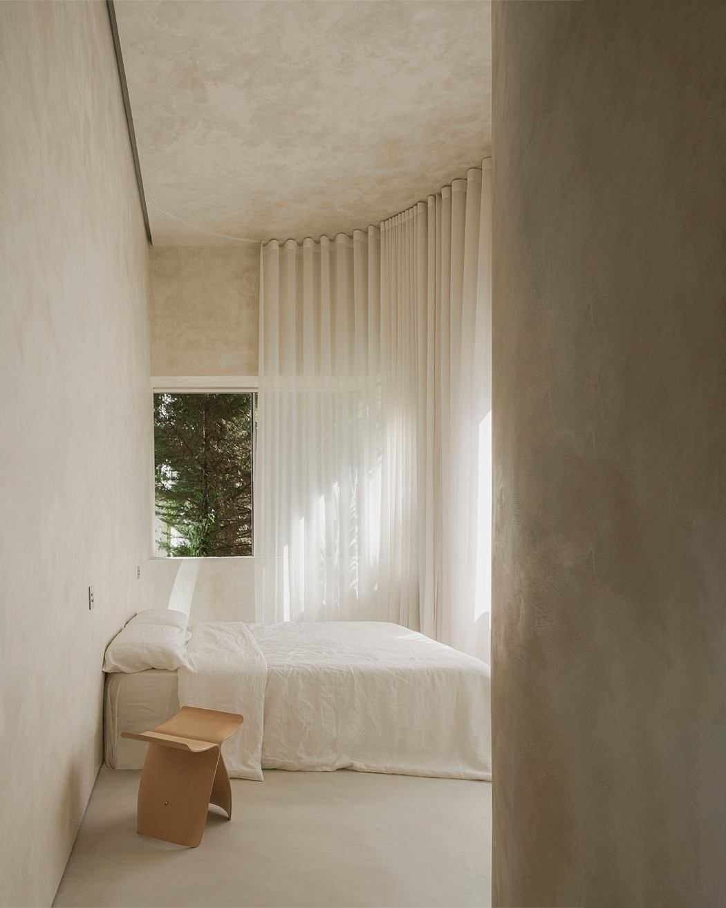 Minimalist bedroom with floor-to-ceiling curtains, wooden side table, and large window overlooking greenery.