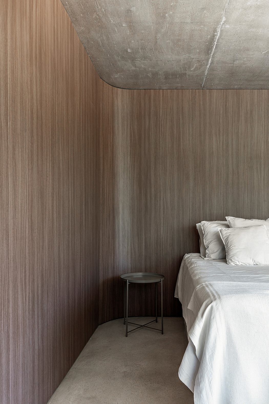 Minimalist bedroom with wood-paneled walls and a concrete ceiling, complemented by a small side table.