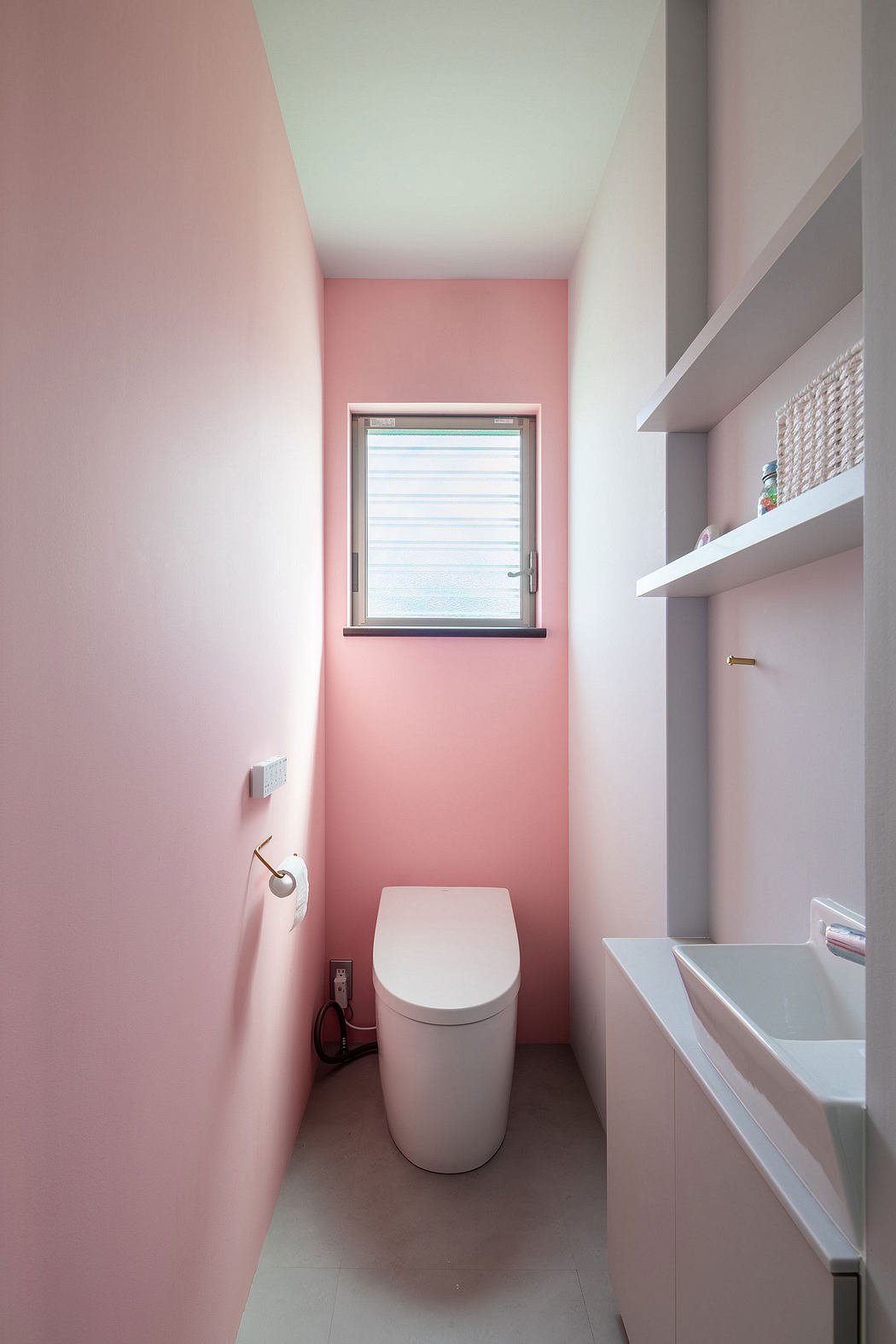 A modern bathroom with a white toilet and vanity, accented by soft pink walls and shelves.