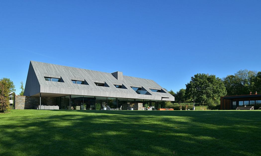 Large, modern building with slate roof and floor-to-ceiling windows, set amidst lush greenery.