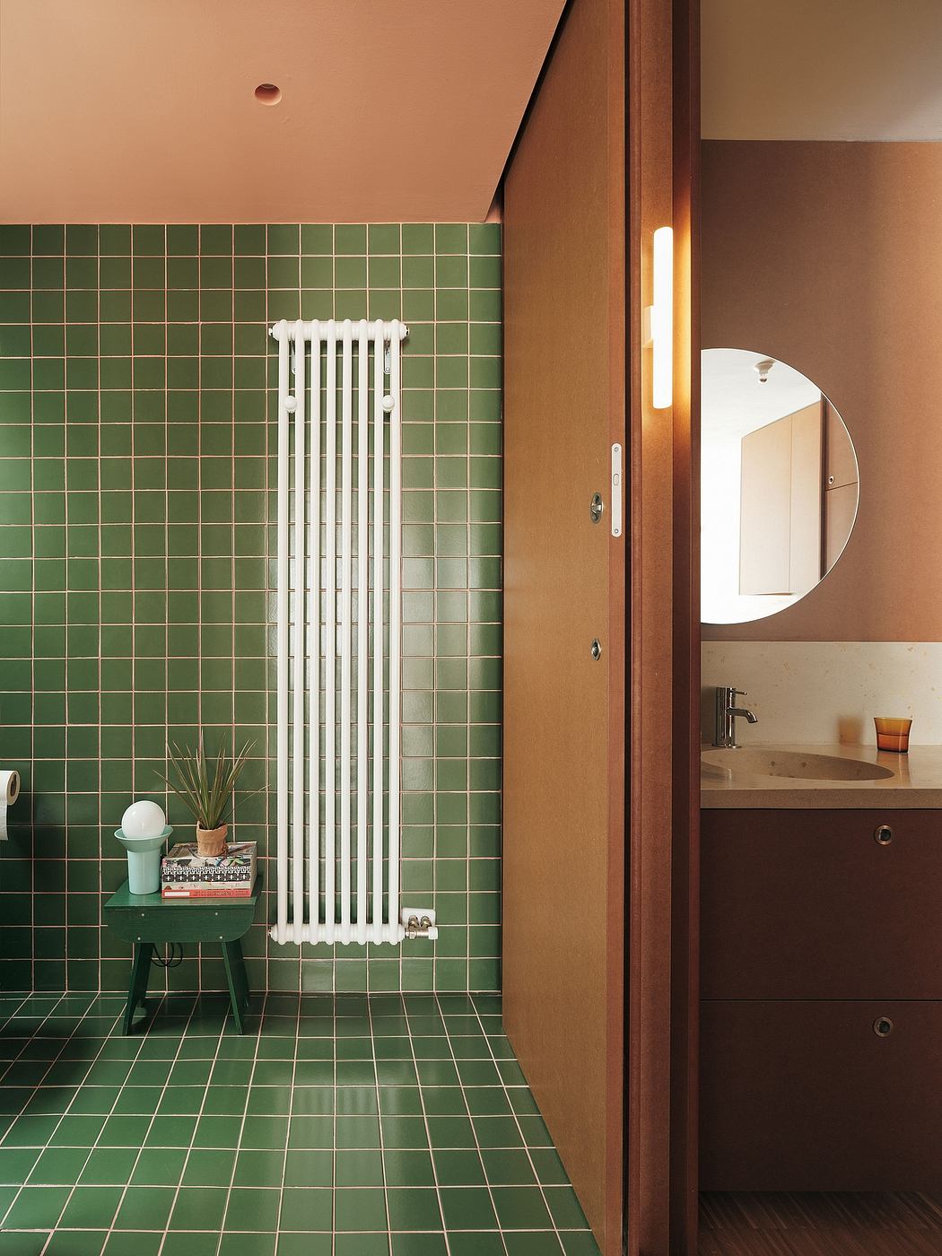 A modern bathroom with a retro-style white radiator, green tile walls, and a wooden vanity.