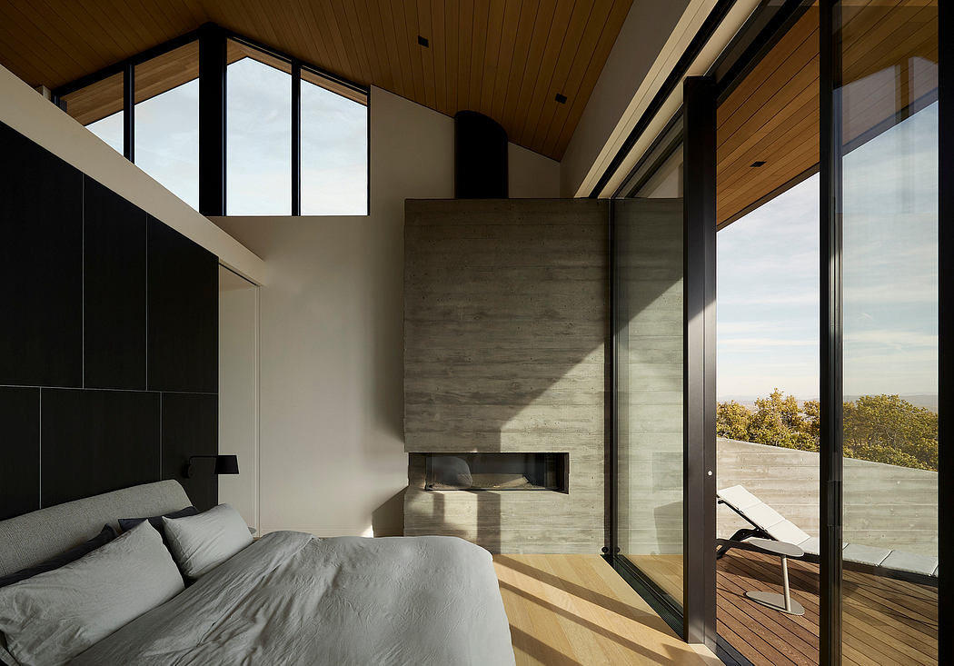 Expansive bedroom with concrete fireplace, wooden beams, and panoramic glass walls.