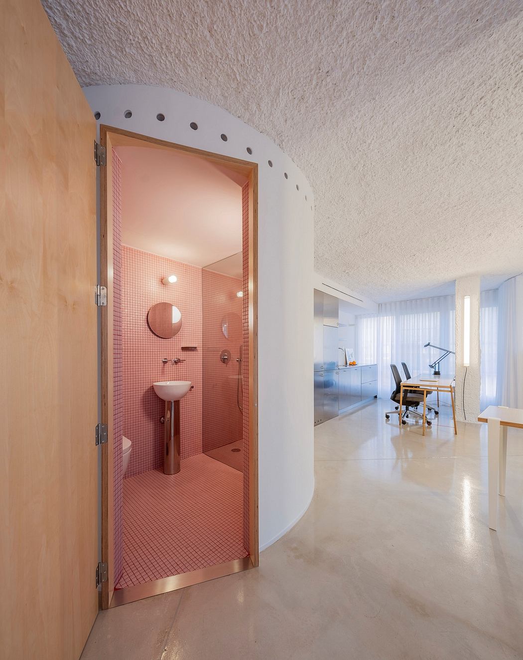 A modern bathroom with pink tile walls and a circular entryway framed by a wooden door.