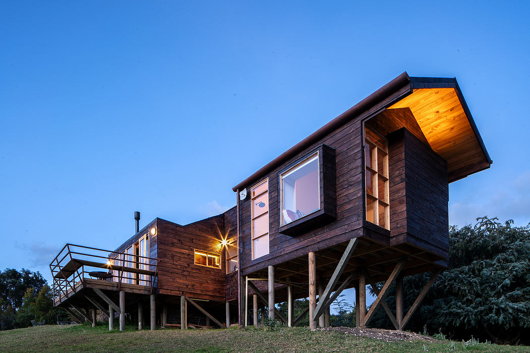 A spacious wooden cabin on stilts with an open balcony and large windows, illuminated at night.