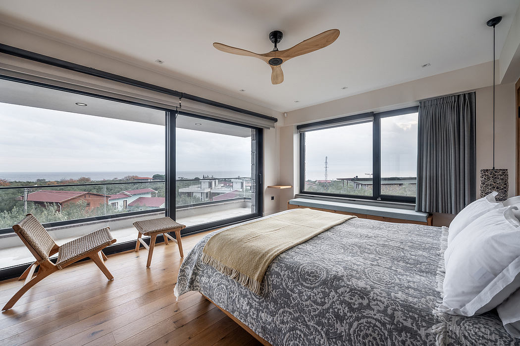 A modern, cozy bedroom with large windows offering a scenic view, featuring a ceiling fan and wooden furniture.