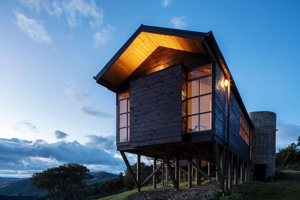 A rustic wooden cabin perched atop stilts, with large windows and a sloping roof.