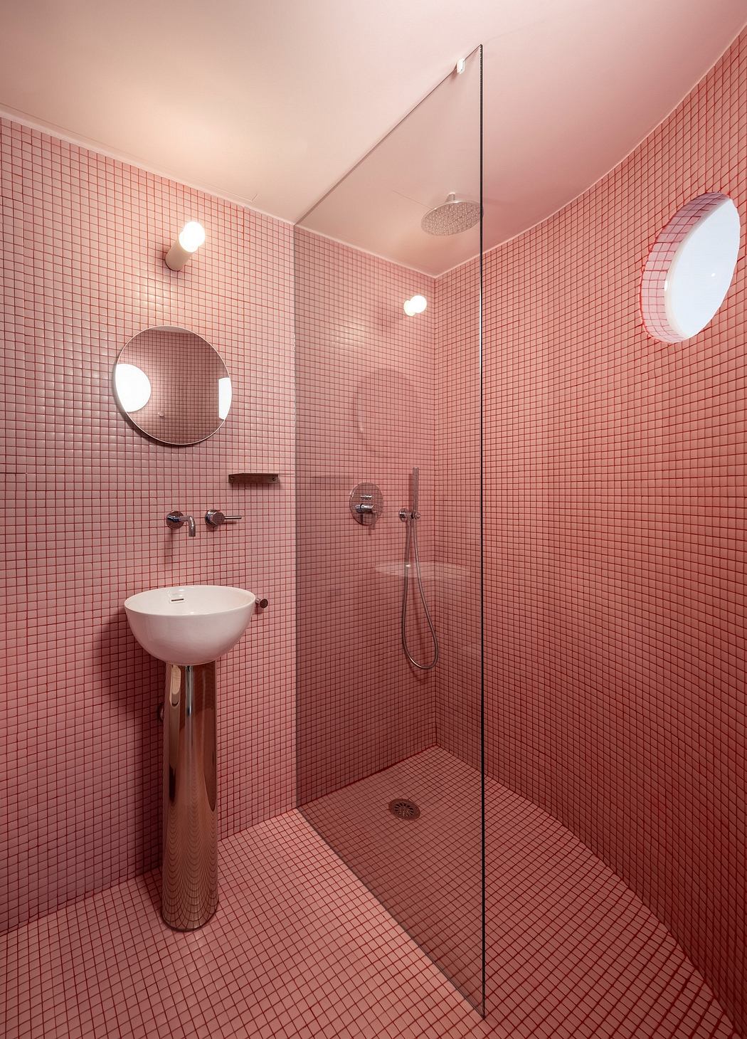 A modern bathroom with pink mosaic tiles, a round mirror, and a glass shower enclosure.