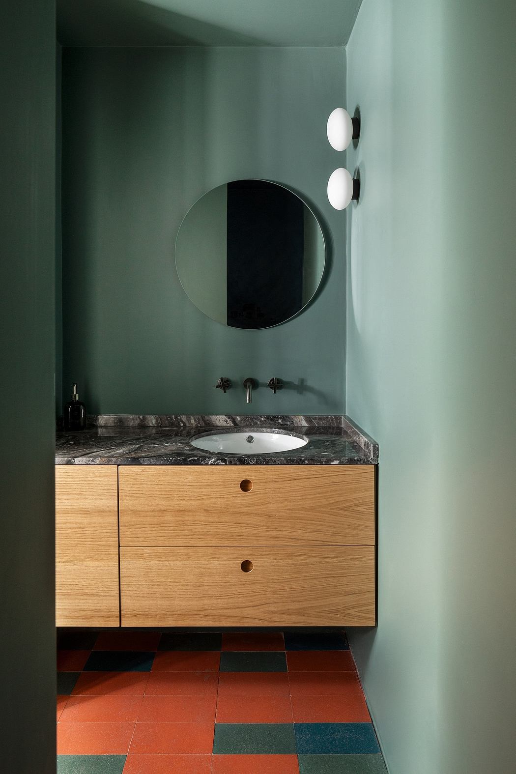 A minimalist bathroom design featuring a round mirror, wooden vanity, and a multicolored tiled floor.