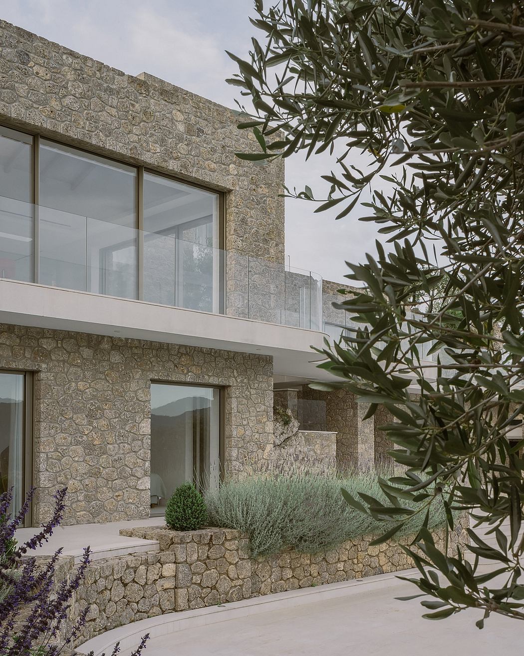 A modern stone building with glass walls and a balcony, surrounded by olive trees.