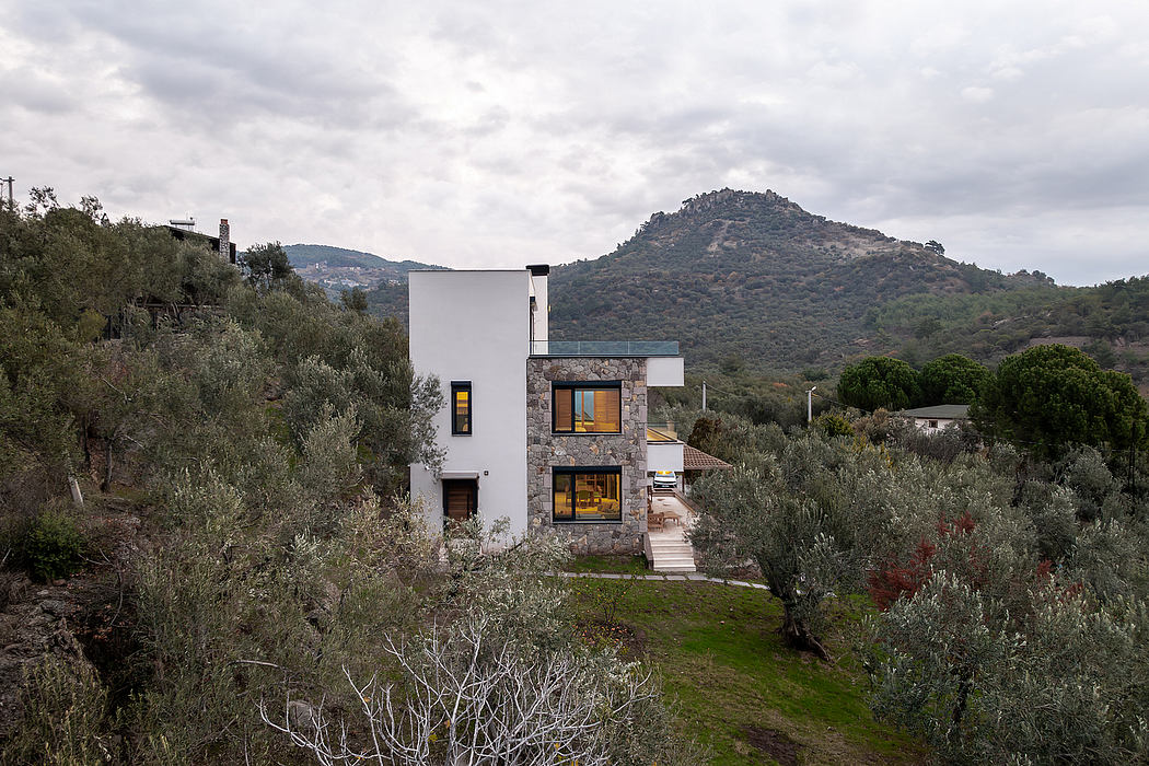A modern, two-story home nestled in a lush, forested landscape with a rugged mountain backdrop.