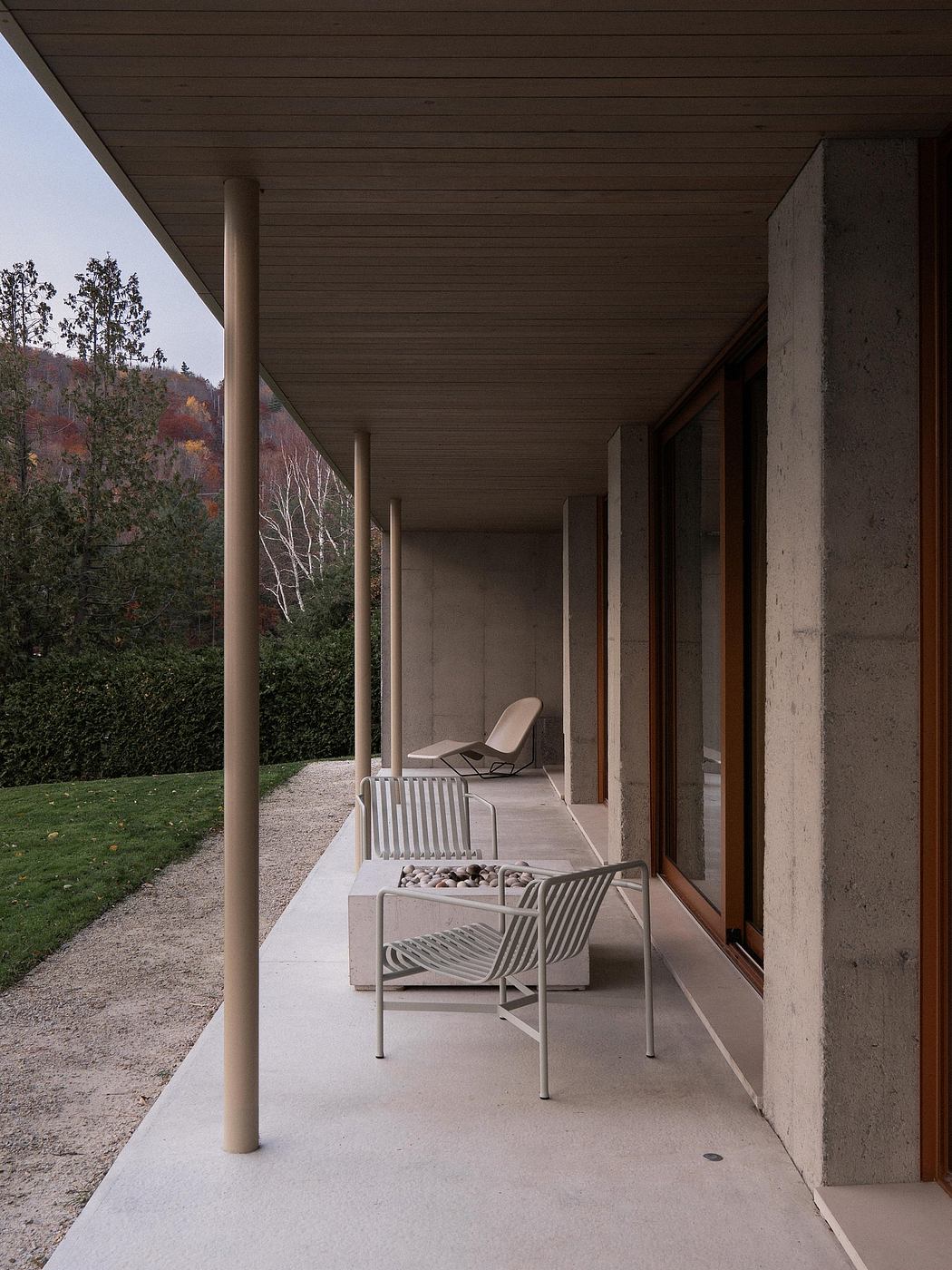 Minimalist concrete porch with wooden ceiling and metal chairs overlooking trees.