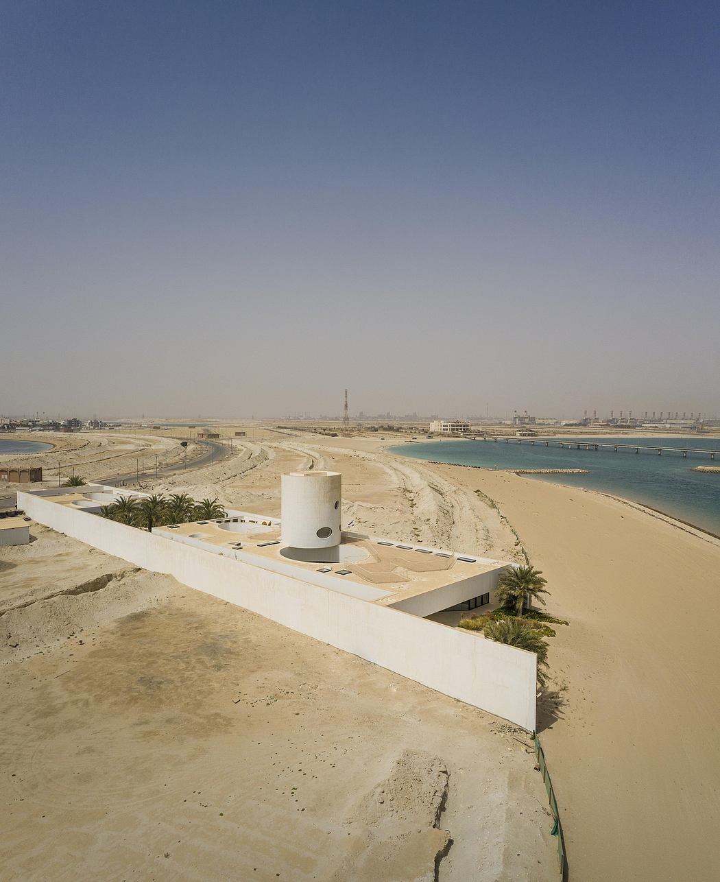Stark desert landscape with white concrete structure and palm trees overlooking water.