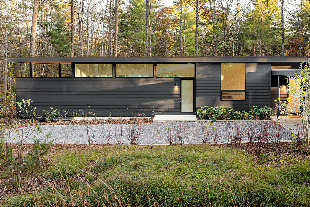 A modern, minimalist cabin nestled in a lush, wooded landscape. Large windows, contrasting dark siding, and natural plantings.